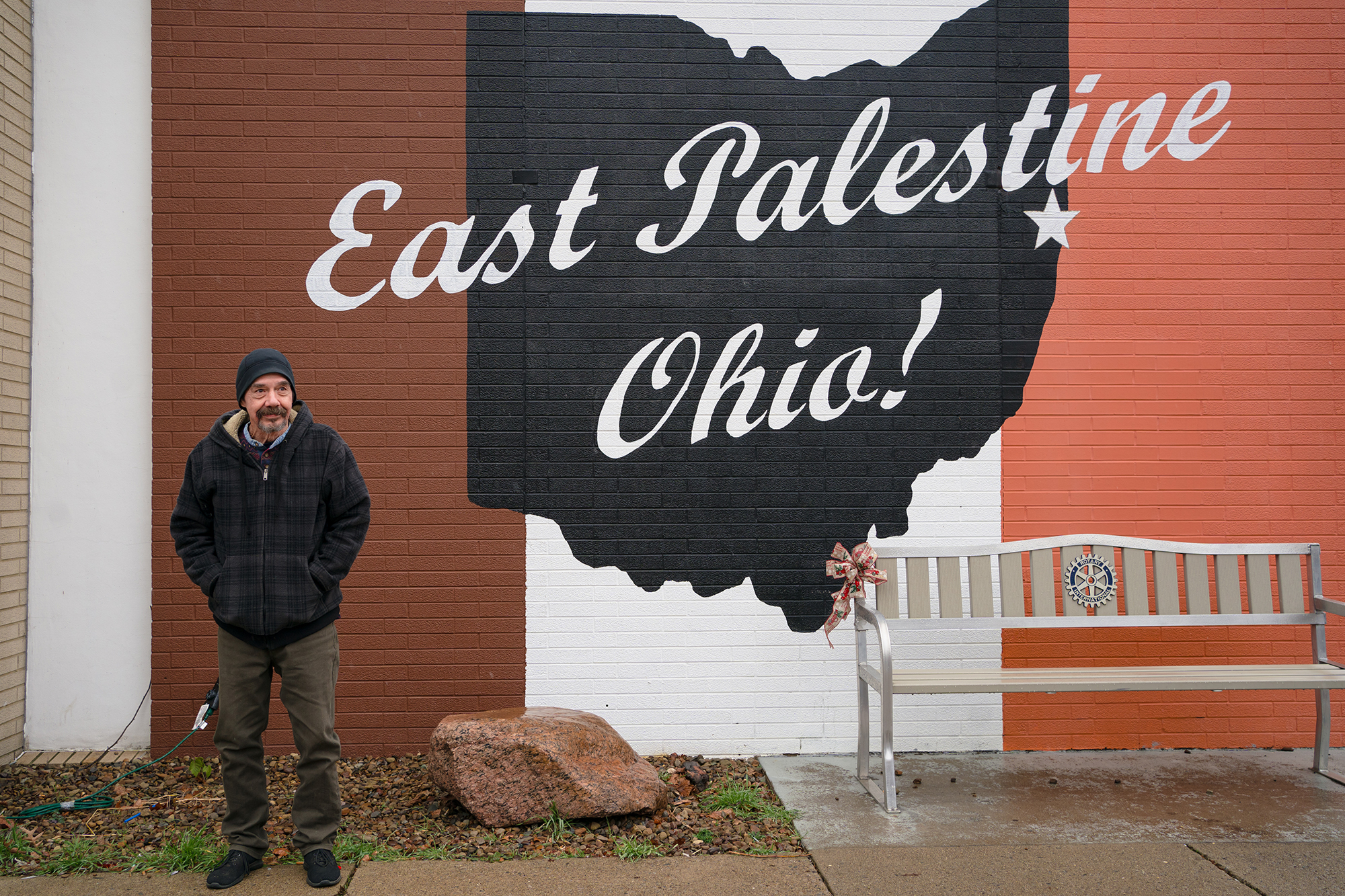 A man wearing a dark winter coat and a knit hat stands in front of a mural that says East Palestine, Ohio.