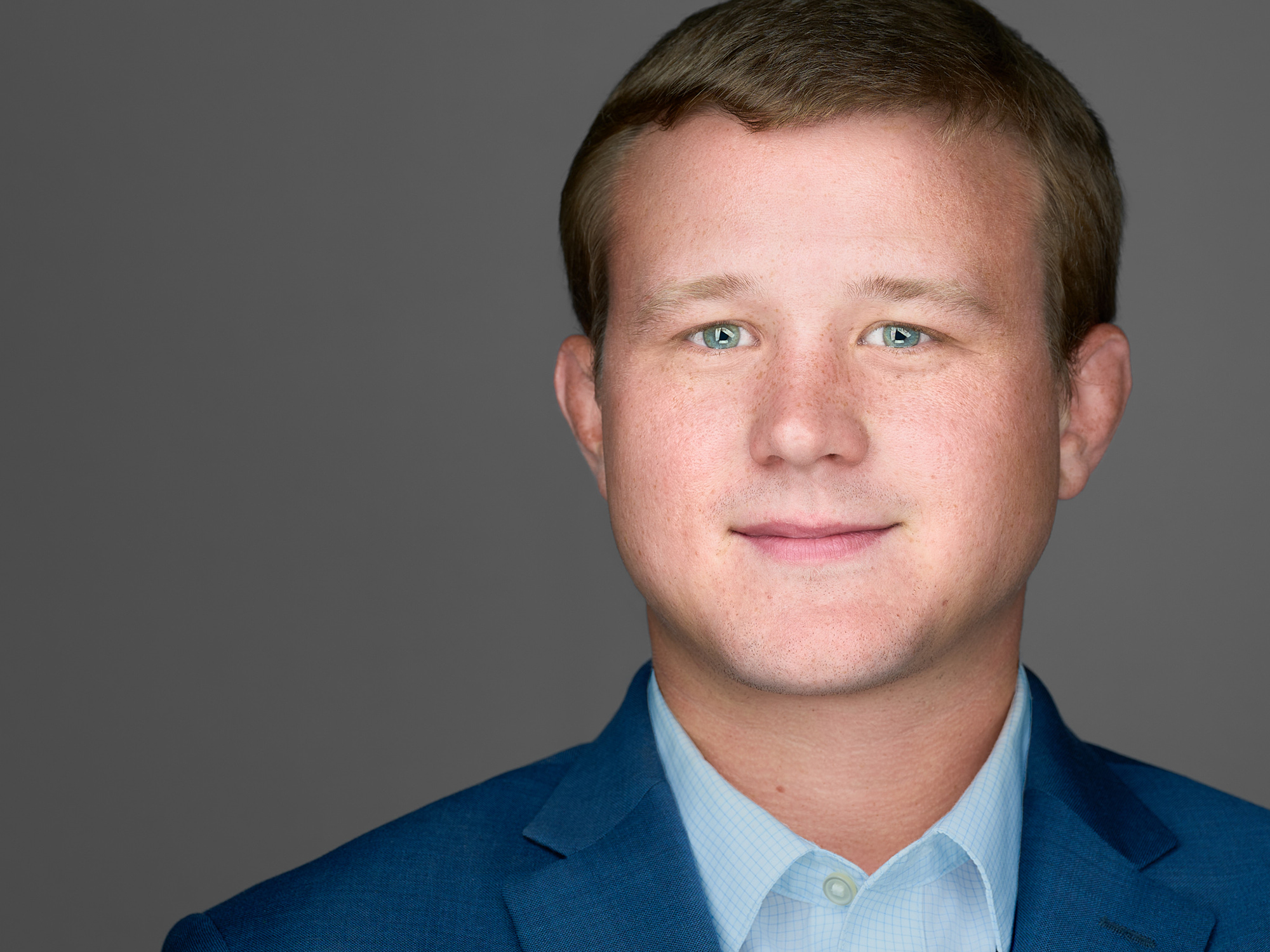 Taylor Nelson poses for a headshot in a blue suit against a grey backdrop.