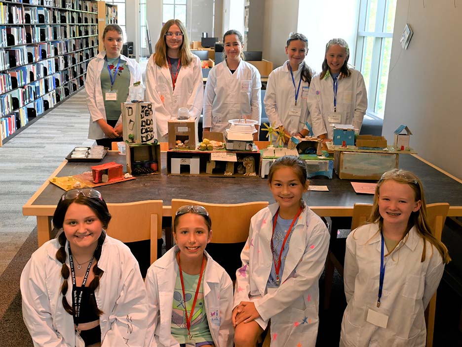 A group of young students in lab coats posing in two rows in front of and behind a table.