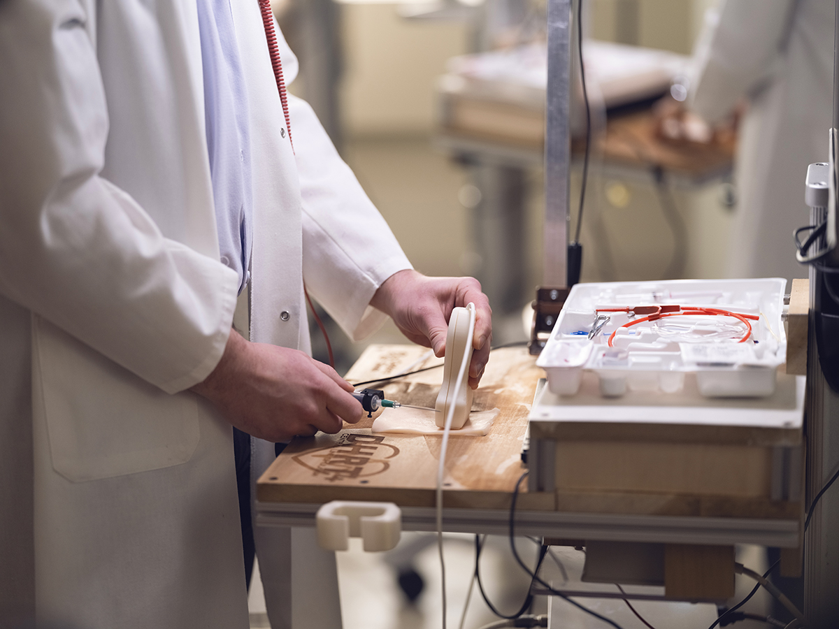 A person in a white coat stands in front of a table holding a needle and an ultrasound wand