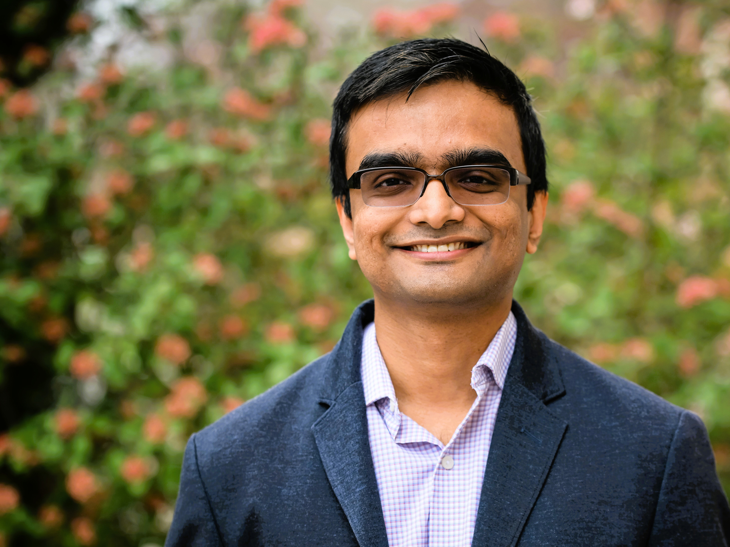 A person wearing glasses and business casual clothing smiles for a photo outside in front of a pink and white-flowered bush.