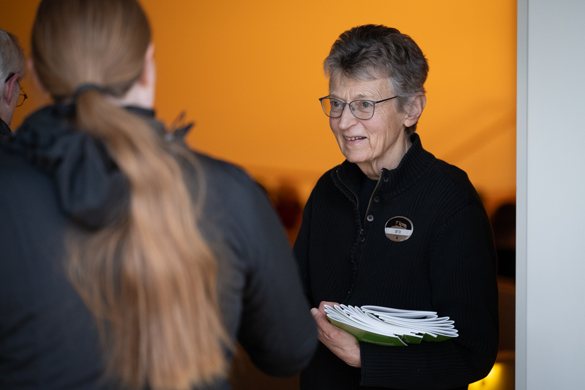 A woman wearing glasses holds a stack of programs that she hands out to patrons entering an auditorium.