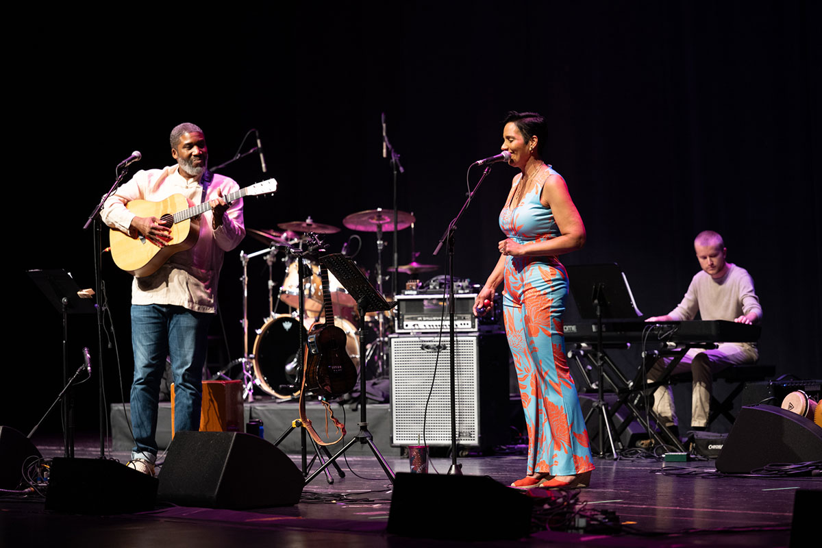 A man of color stands and plays an acoustic guitar while a woman stands and sings near a microphone.