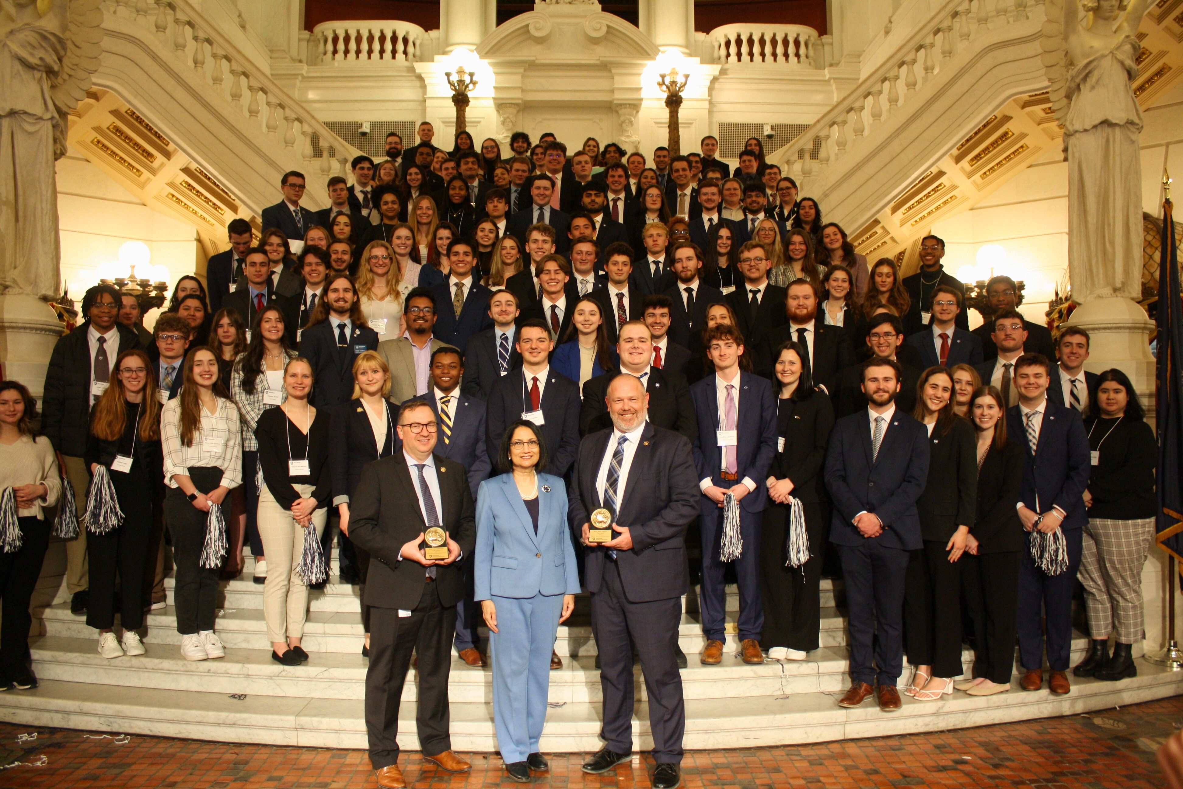 one woman and two men standing in front of a large group of students on steps in the Pennsylvania state capitol building. 