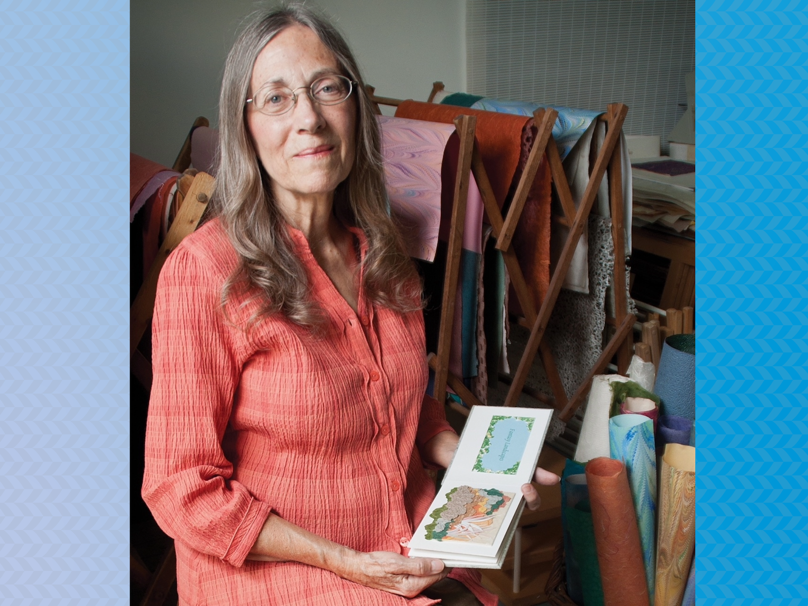 photo portrait of woman with glasses holding small wide artist book near drying rack holding colorful sheets of art paper and tall standing art paper rolls