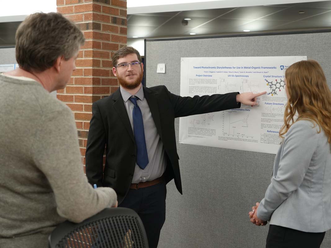 Student in a suit gesturing to a white poster board as others look on.