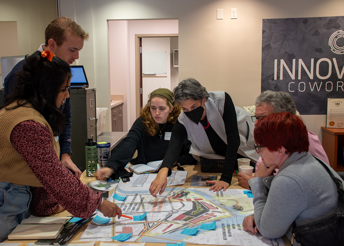 Lisa Iulo leans across a table of plans at a meeting with community members and students surrounding the table.