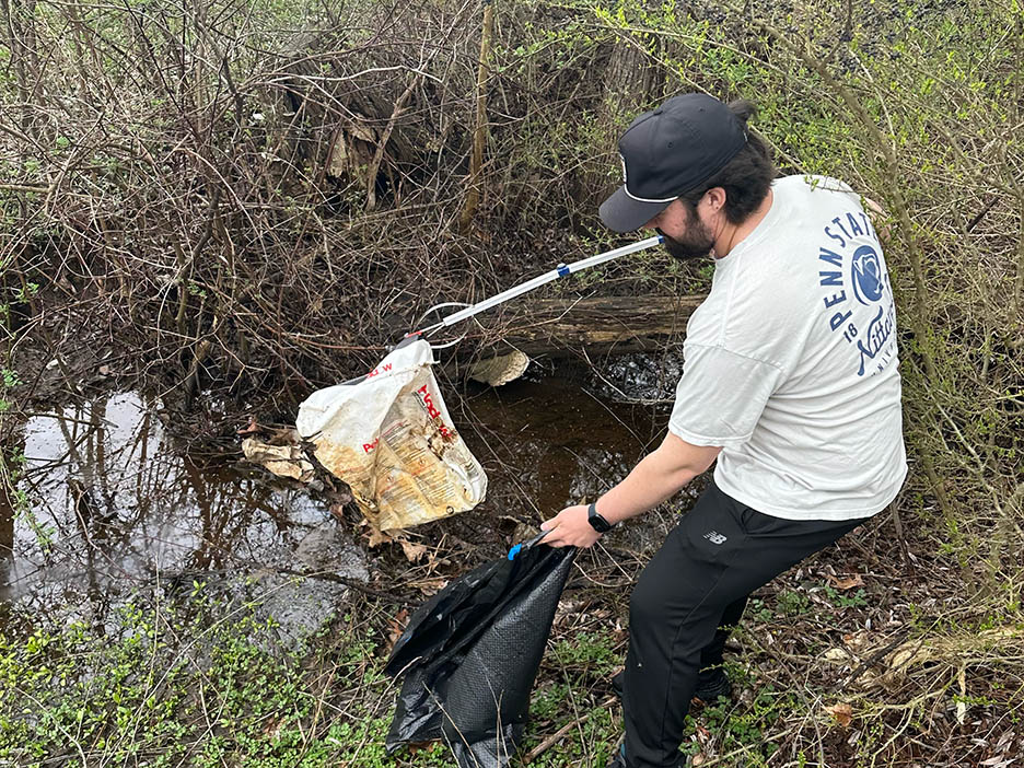 A person holds a garbage bag open with one hand while picking up garbage with a tool in the other.