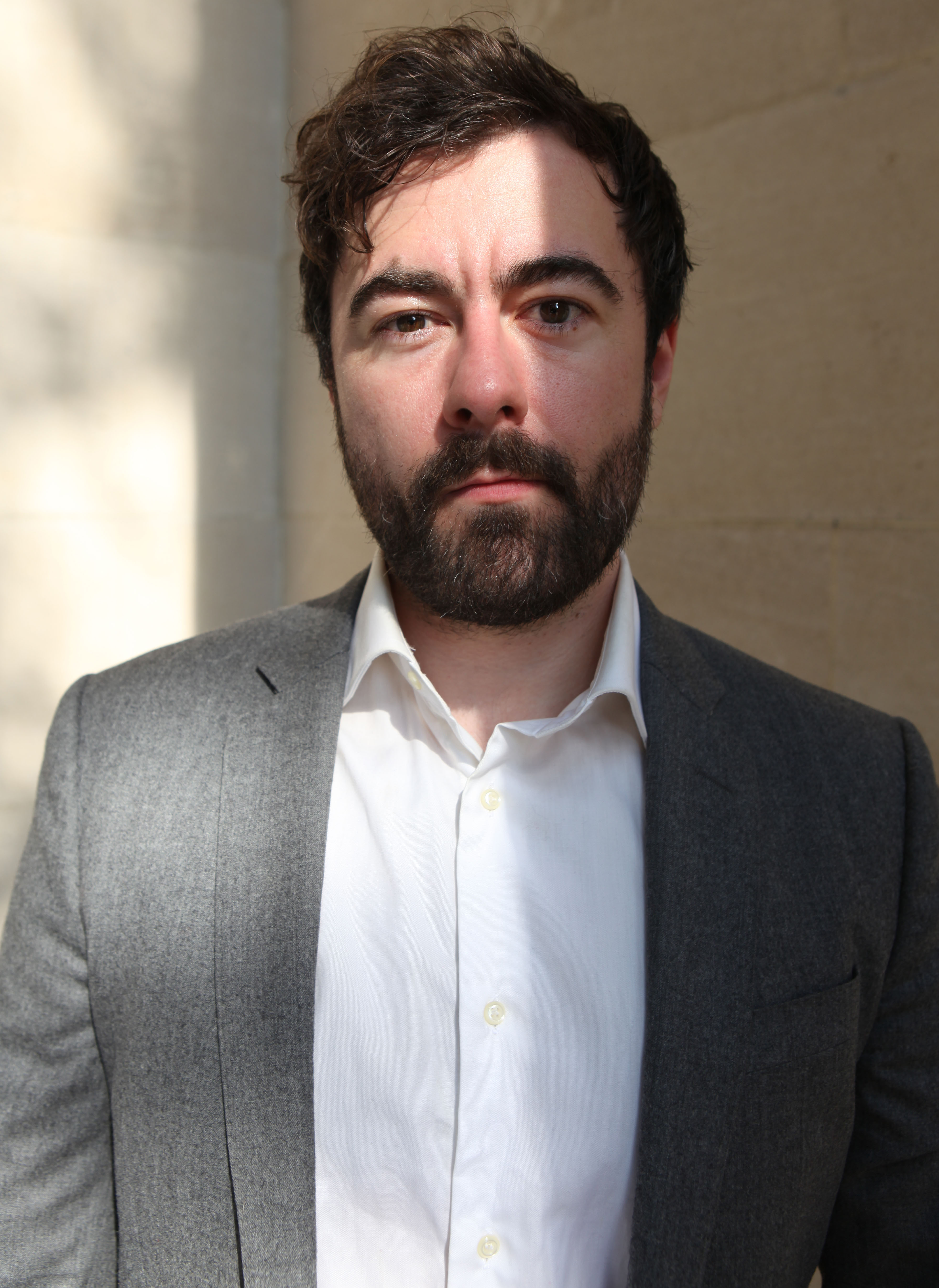 White man with dark hair posing with a grey blazer and white shirt