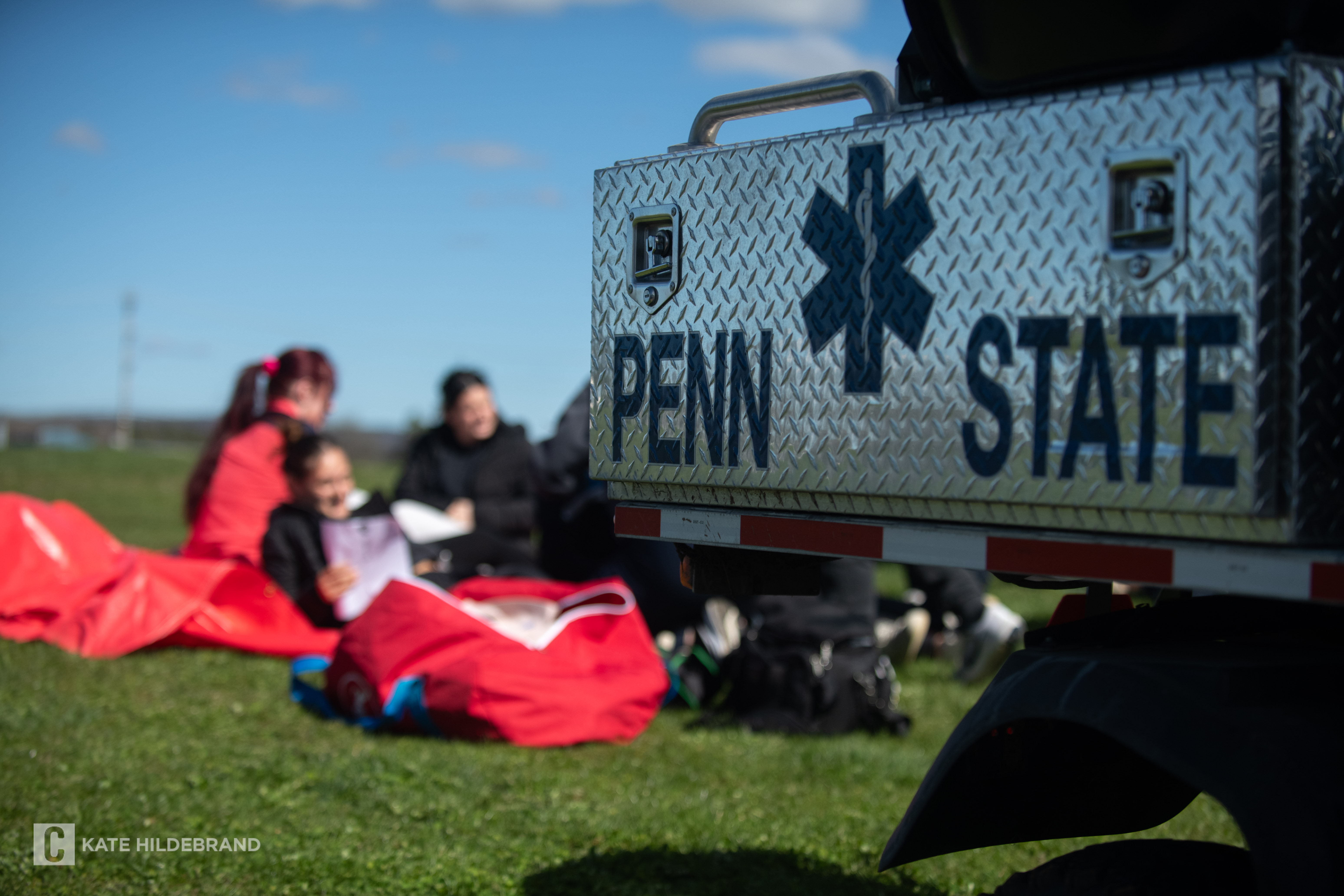 A Penn State emergency vehicle is parked on a grassy field with students sitting on the ground in the background.