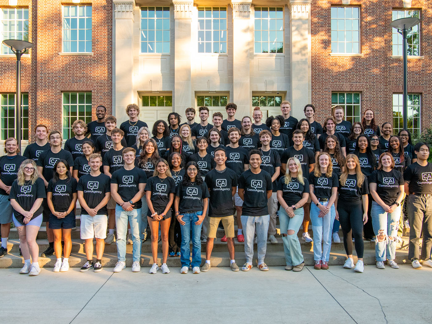 Studnets in black T-shirt with a white logo stand in a large group on the steps of a red brick buiilding