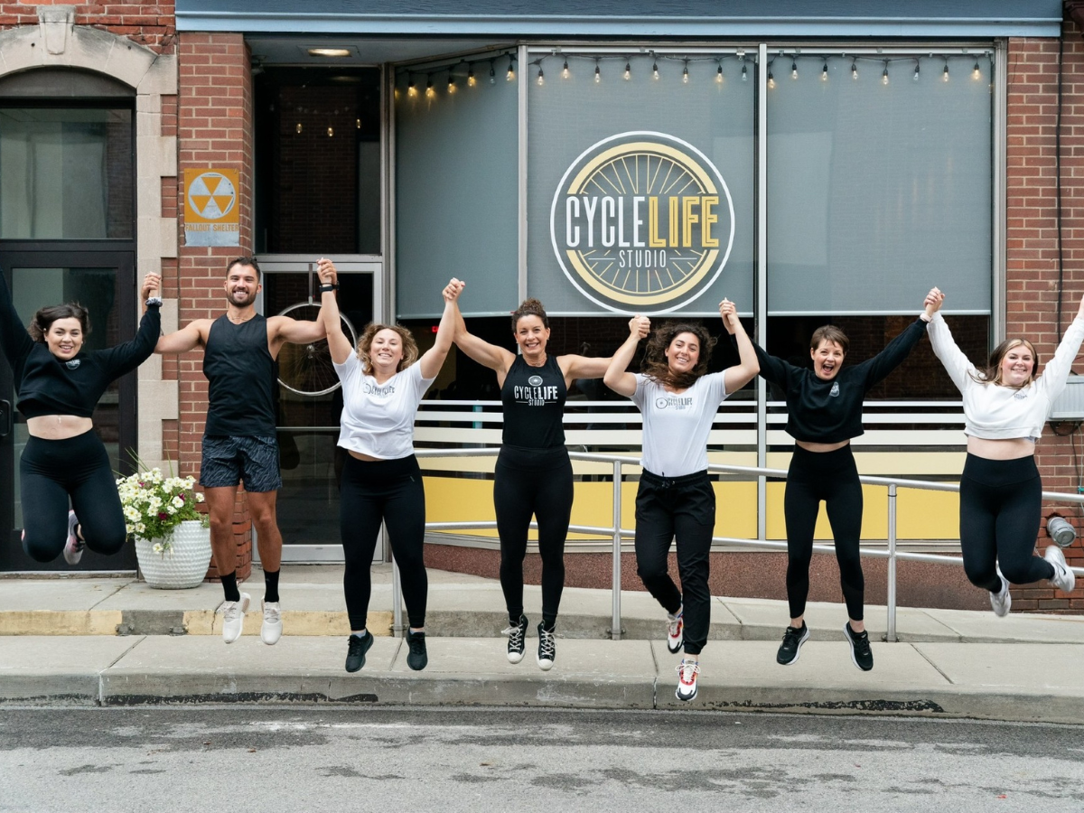 The CycleLife Studio team holds hands and jumps in unison on the street outside of the CycleLife Studio location