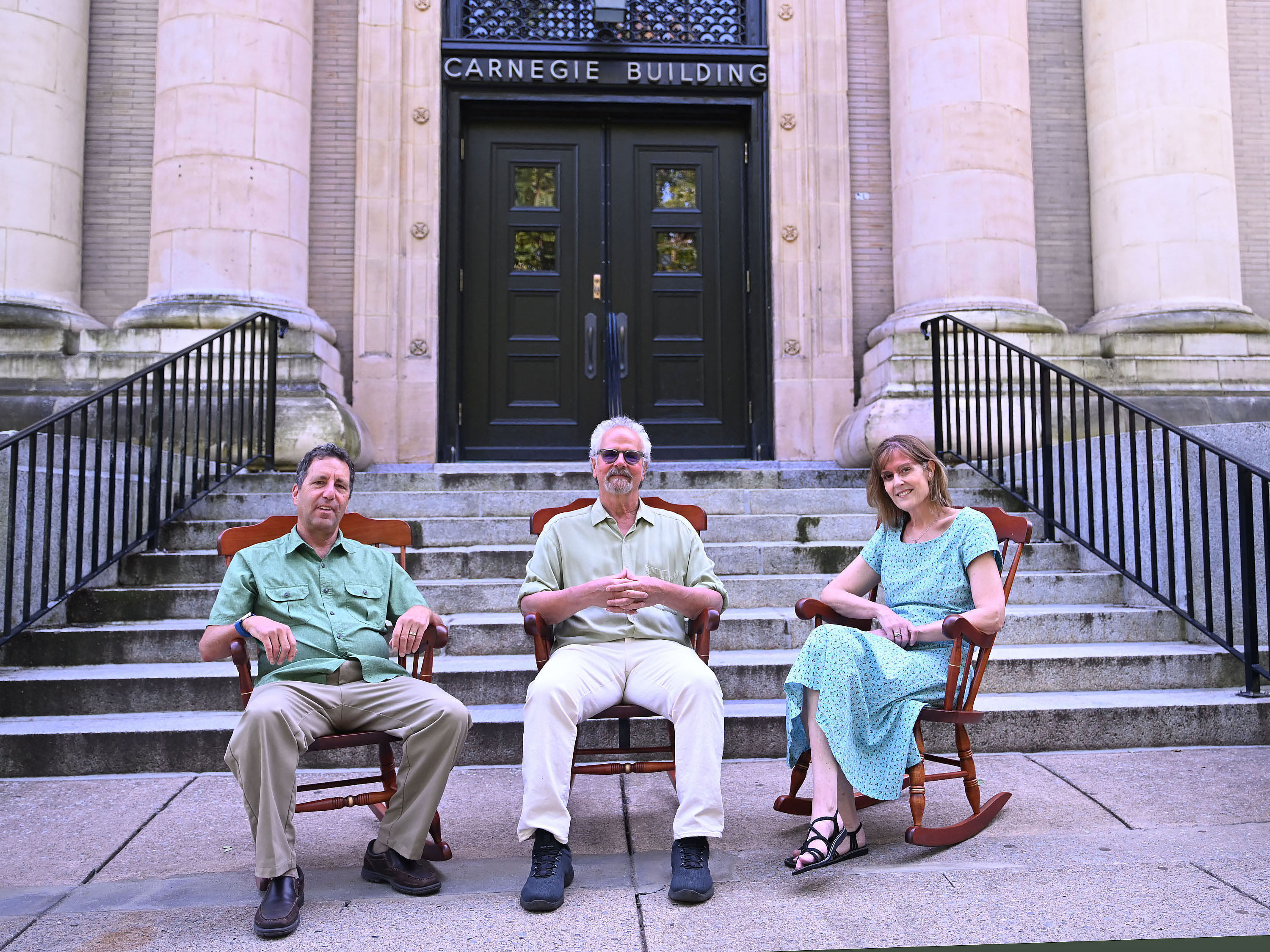 Three people, two men and a woman, sit in rocking chairs in front of a light colored brick building.