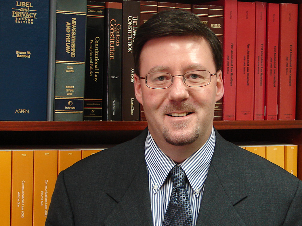 A smiling dark haired main wearing glasses and a coat and tie in front of a bookshelf