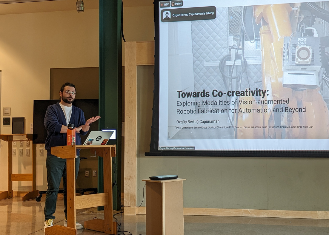 Özgüç Bertuğ Çapunaman stands at a podium delivering his thesis defense.