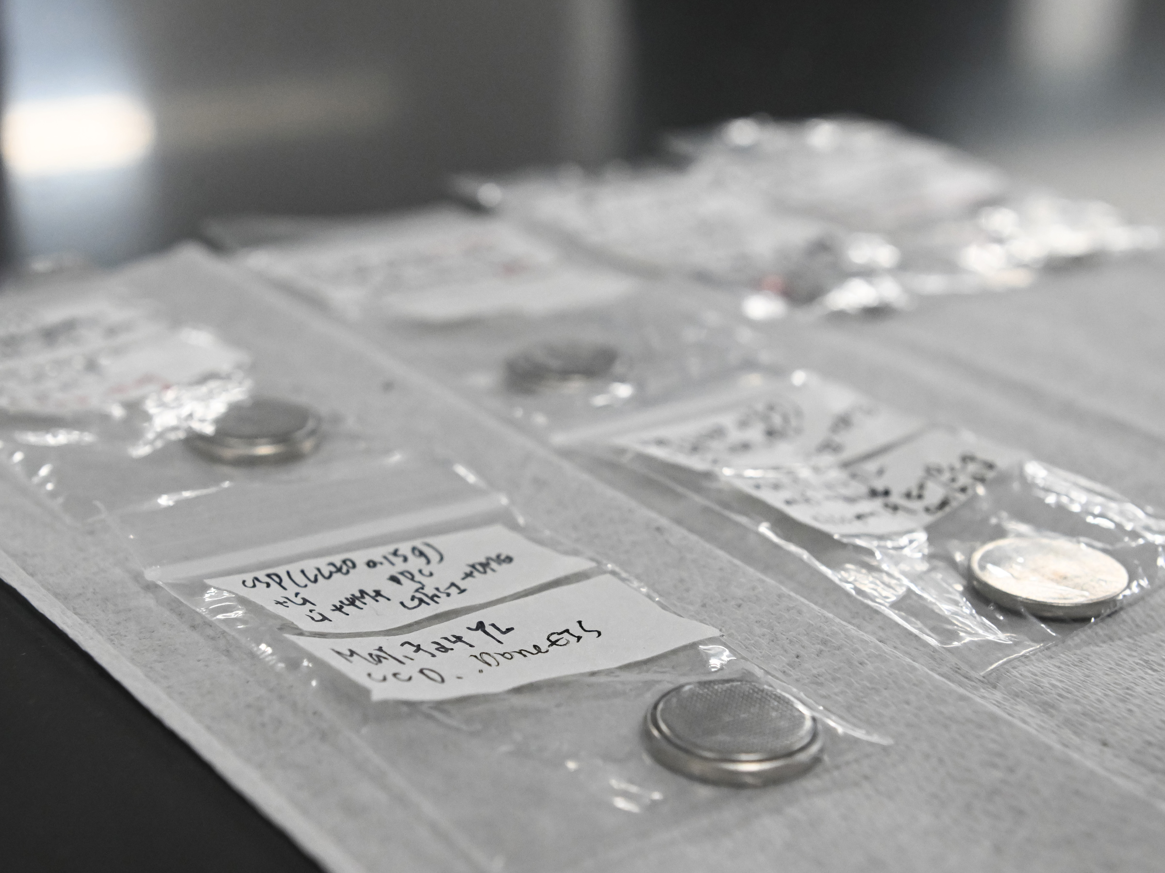 Coin cell battery samples are arranged in plastic bags on a lab table.