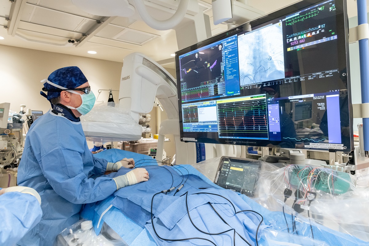 A physician in an operating room wearing scrubs and glasses stands over a patient and looks at a computer monitor.