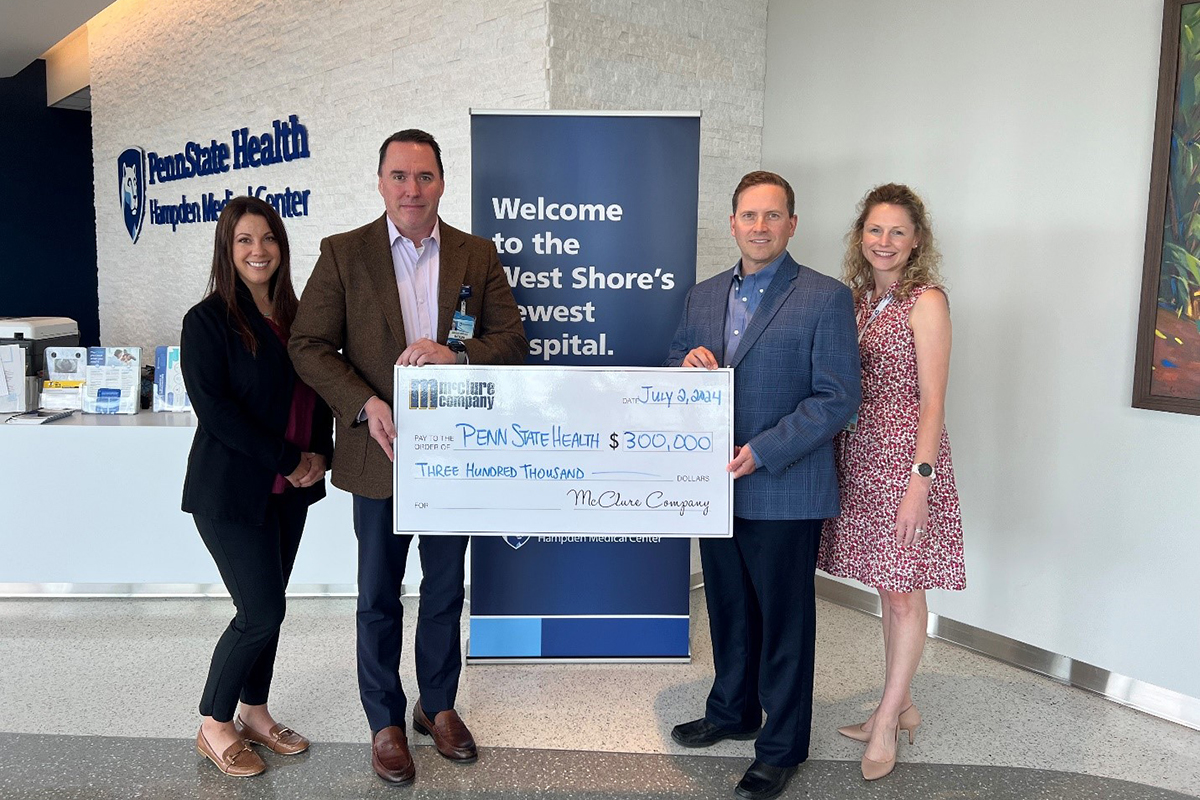 Two men and two women pose in front of a vertical banner that says, “Welcome to the West Shore’s newest hospital.” The gentlemen are in the middle holding a large presentation check for $300,000 written to Penn State Health from McClure Company.