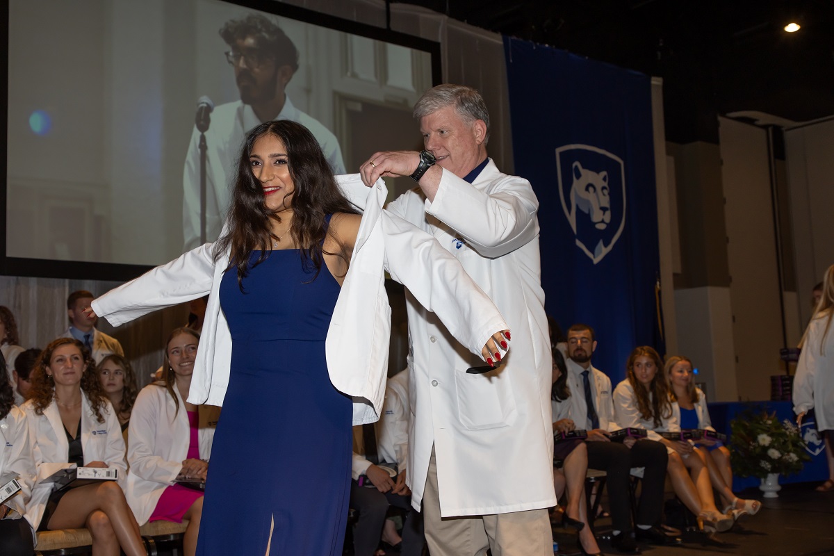 A man wearing a white doctor's coat presents a young woman with her own doctor's coat at the Penn State College of Medicine White Coat Ceremony