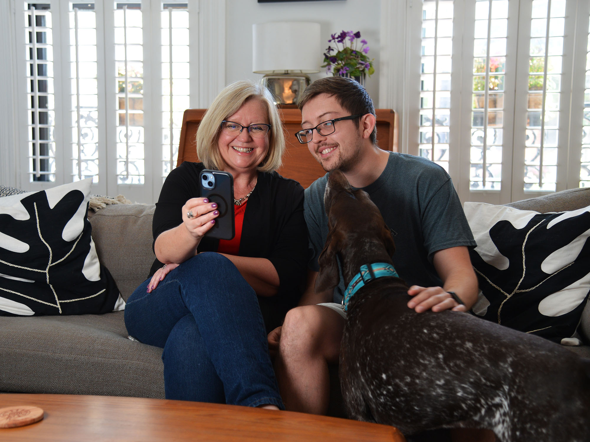 a woman holding a phone sits next to her son on a couch while the son pets a dog