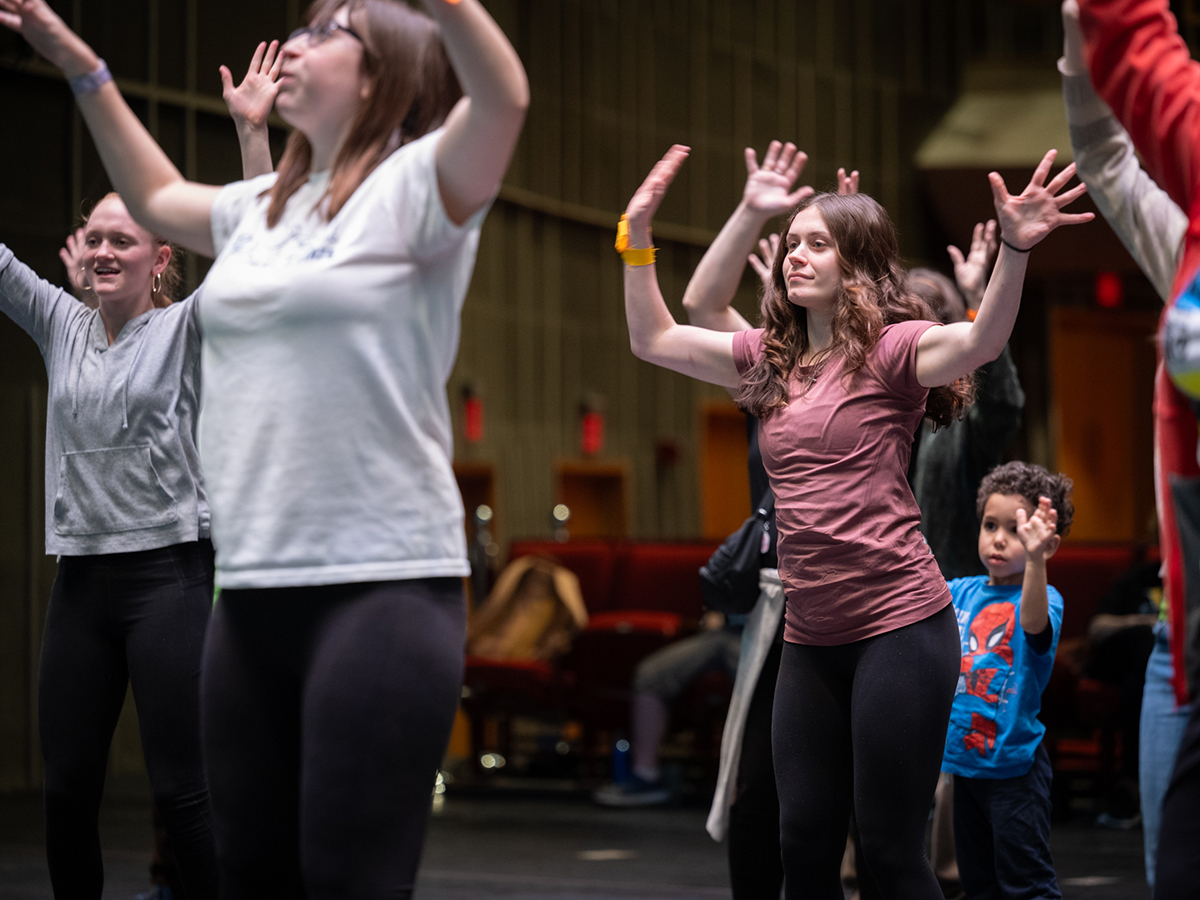 A group of young people dance on a stage. One girl is in focus with her hands waving in the air