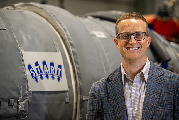 A person in glasses smiles at the camera in front of a piece of equipment with a sign that says START. 