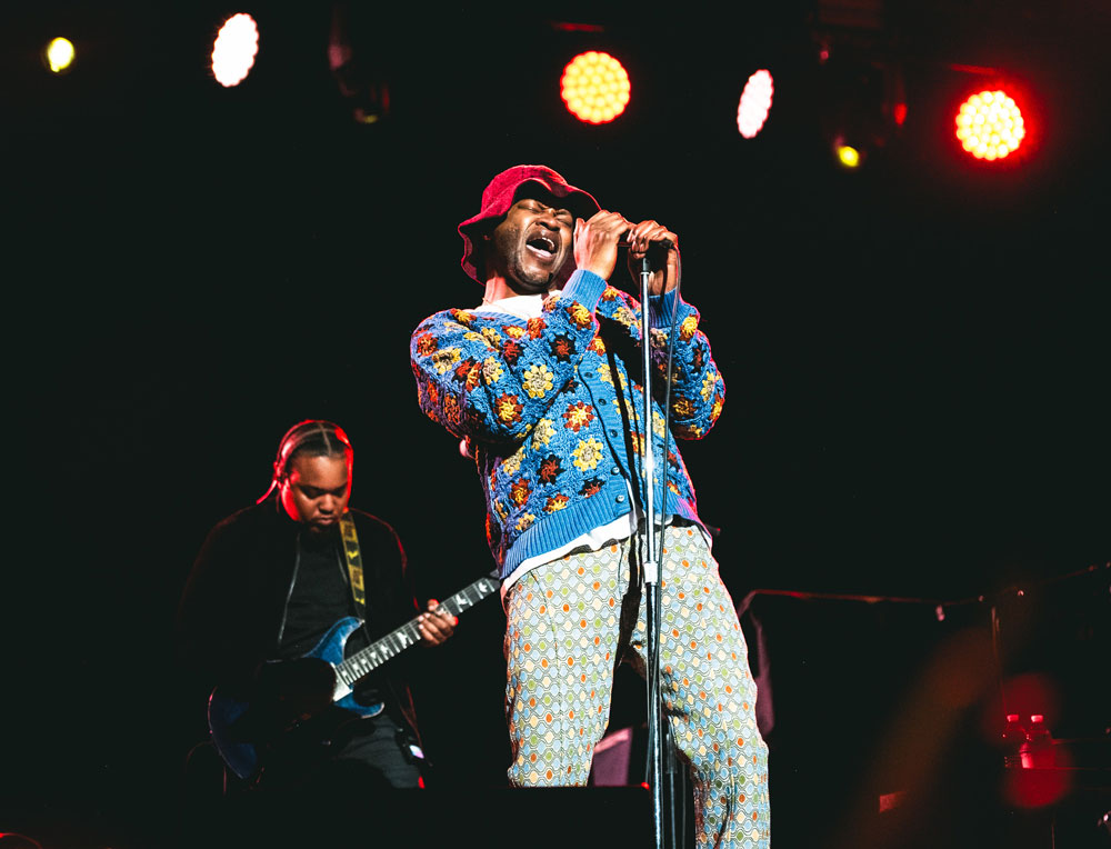 A man wearing a crocheted sweater and pants holds a microphone with both hands as he sings into it.