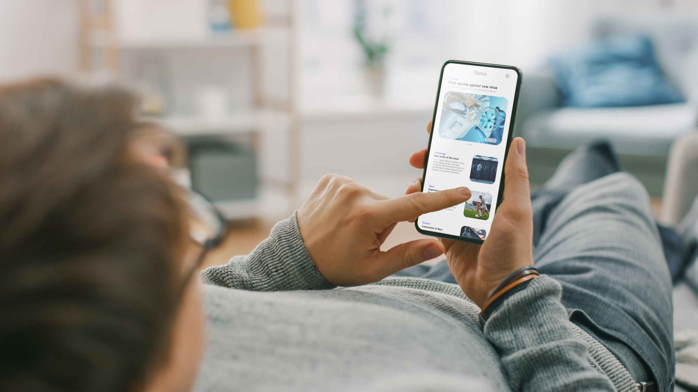 A man on a couch scrolling through news articles on a smartphone