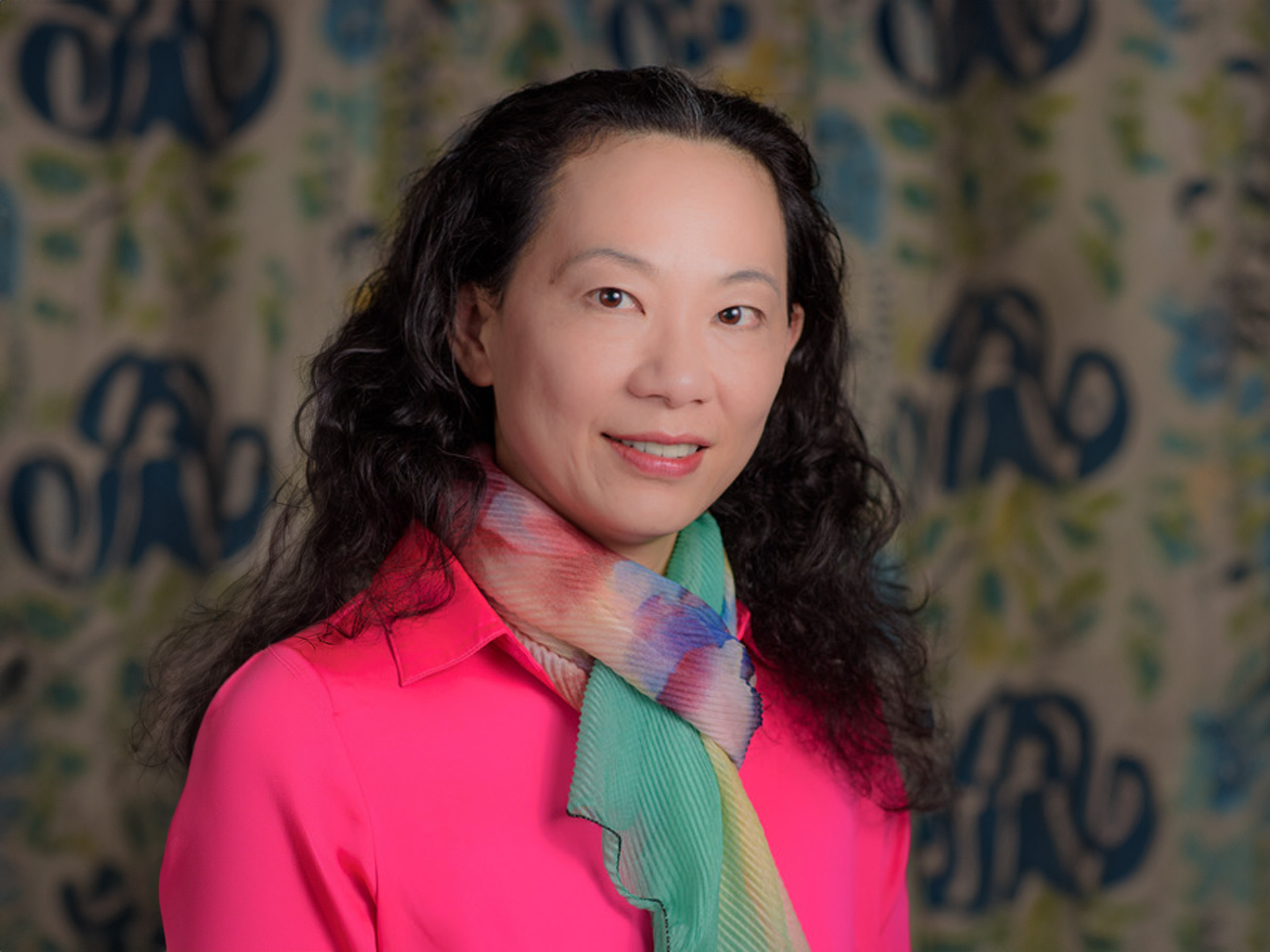 Woman wearing a pink shirt and a colorful scarf, stands in front of a patterned background