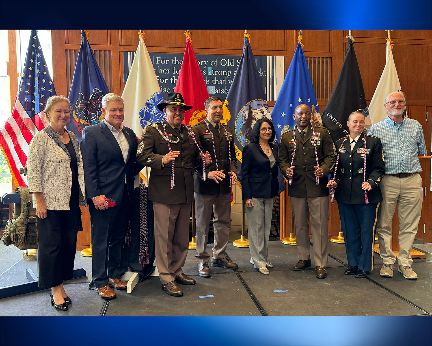 A group of people pose in front of the American flag and U.S. military flags.