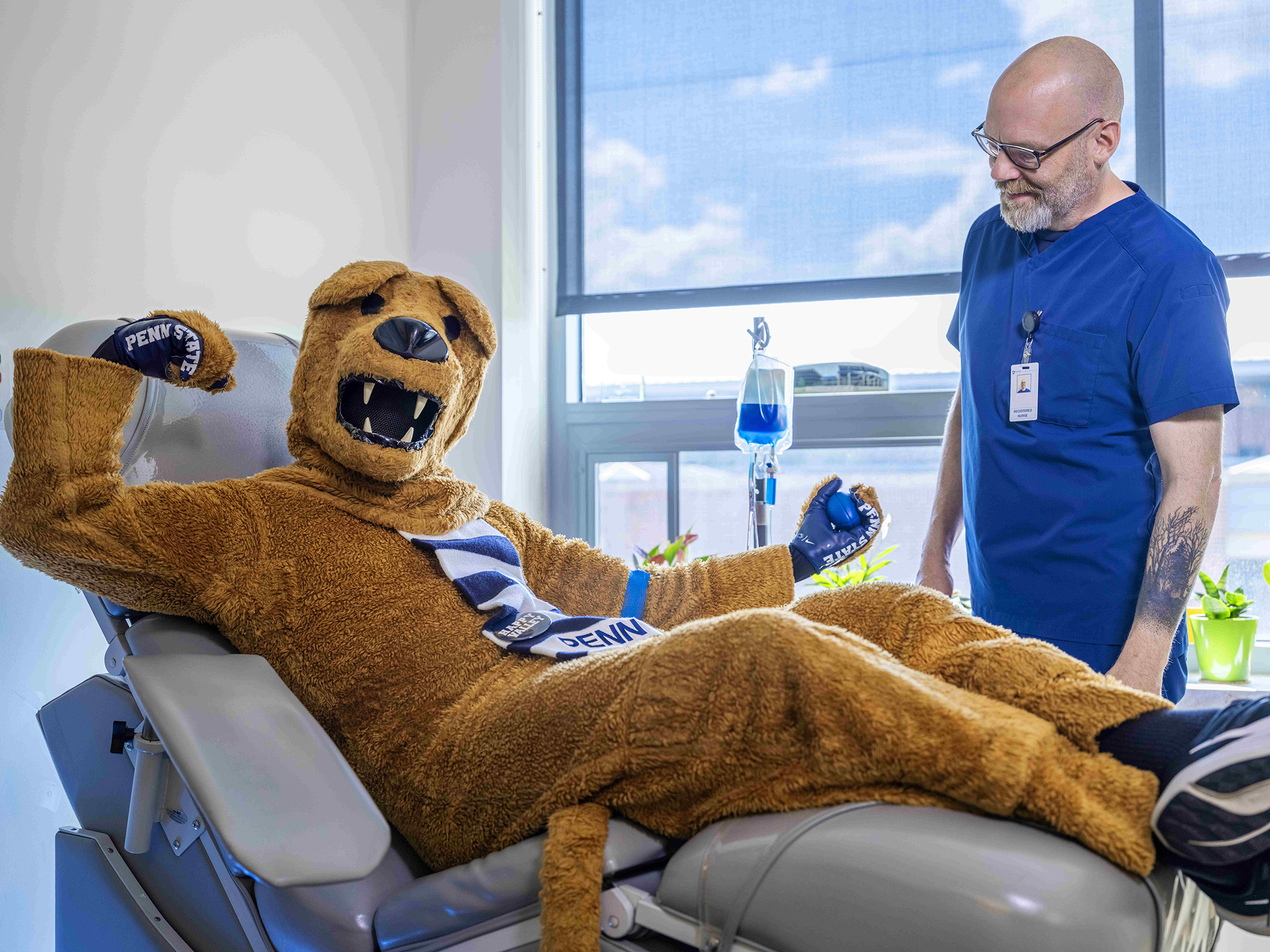 The Nittany Lion sits on a medical bed flexing his arm after donating blood with a smiling attendant nearby