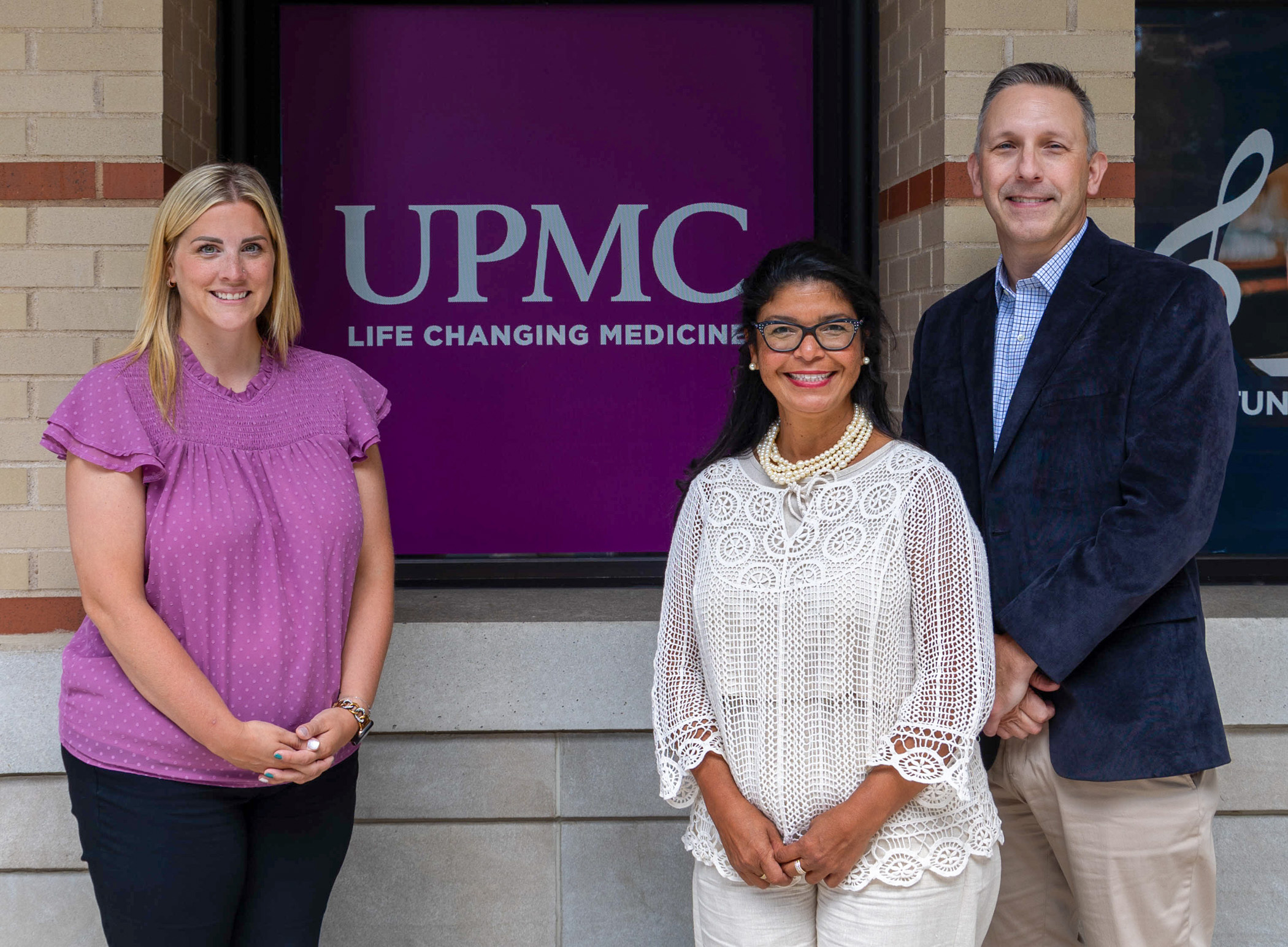 Three people pose for a photo in front of a sign that says UPMC