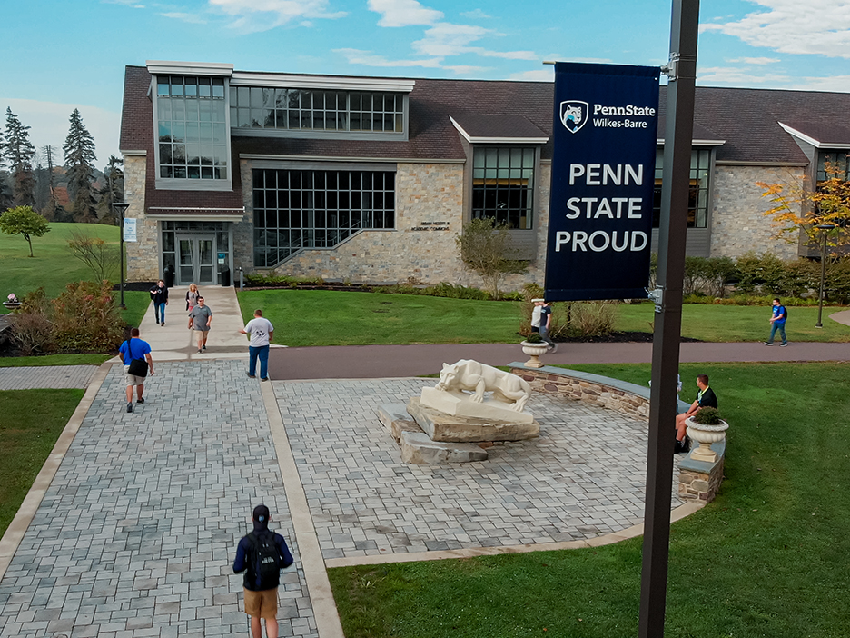 A view looking down at a campus building with large windows, a walkway with pavers and a Nittany Lion statue, with students walking throughout the photo.