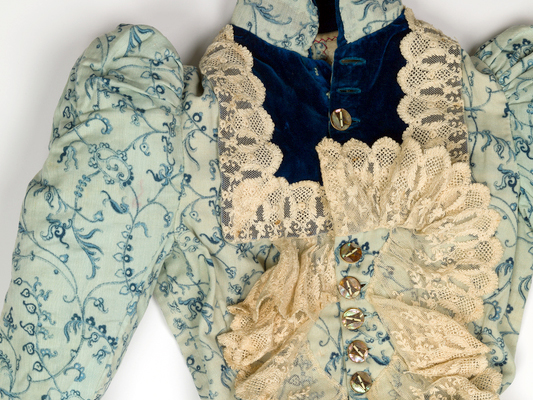 A close-up photo of a vintage light blue-green dress with puffed sleeves, brass buttons and a yellowed, ruffled lace collar