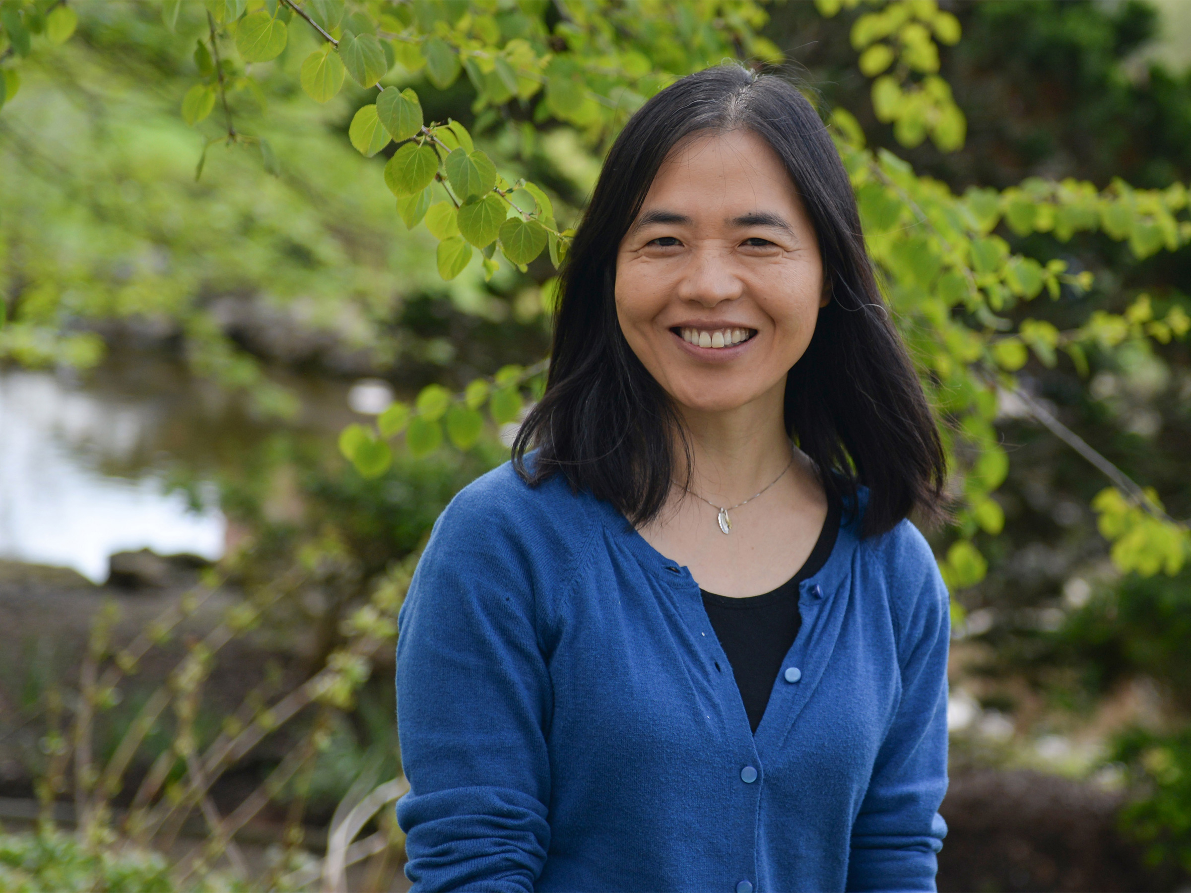 An individual wearing a blue sweater in front of greenery. 