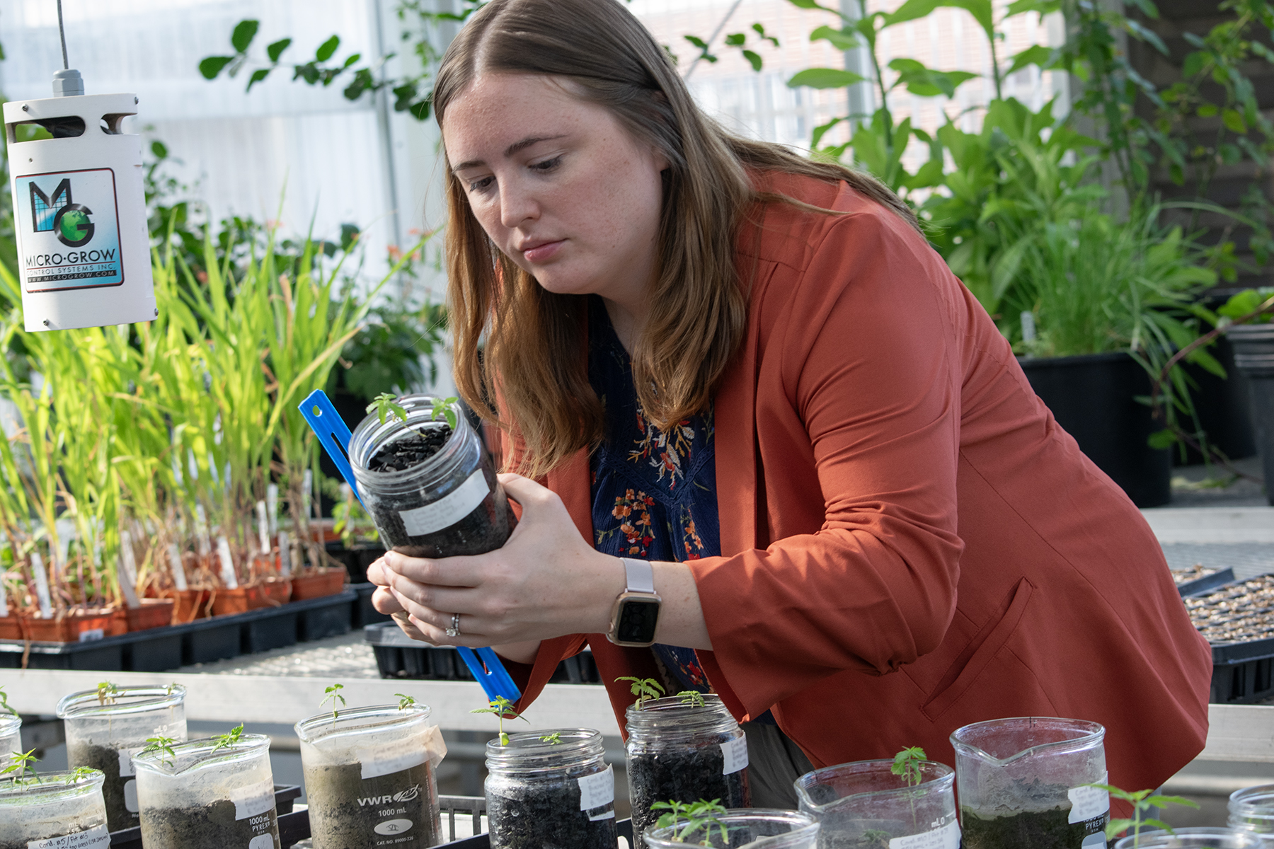 A woman holds a jar of soil and a ruler, taking a measurement, with plants behind her