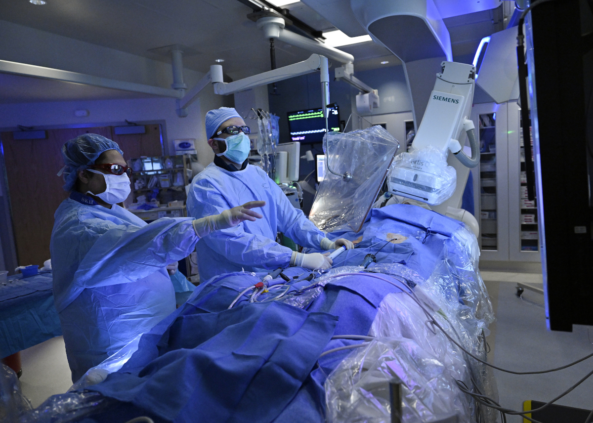 Two physicians perform a procedure on a draped patient in a surgical setting. Various pieces of medical equipment are located throughout the room.