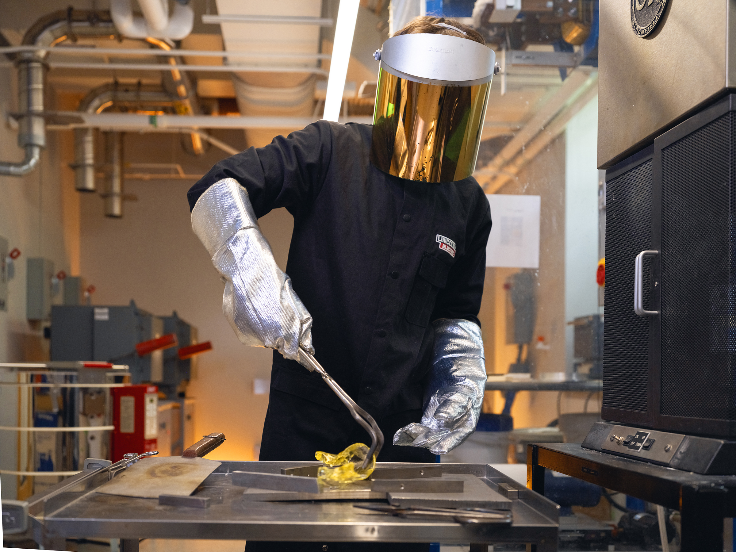 A person wearing a face heat shield molds glass from a forge