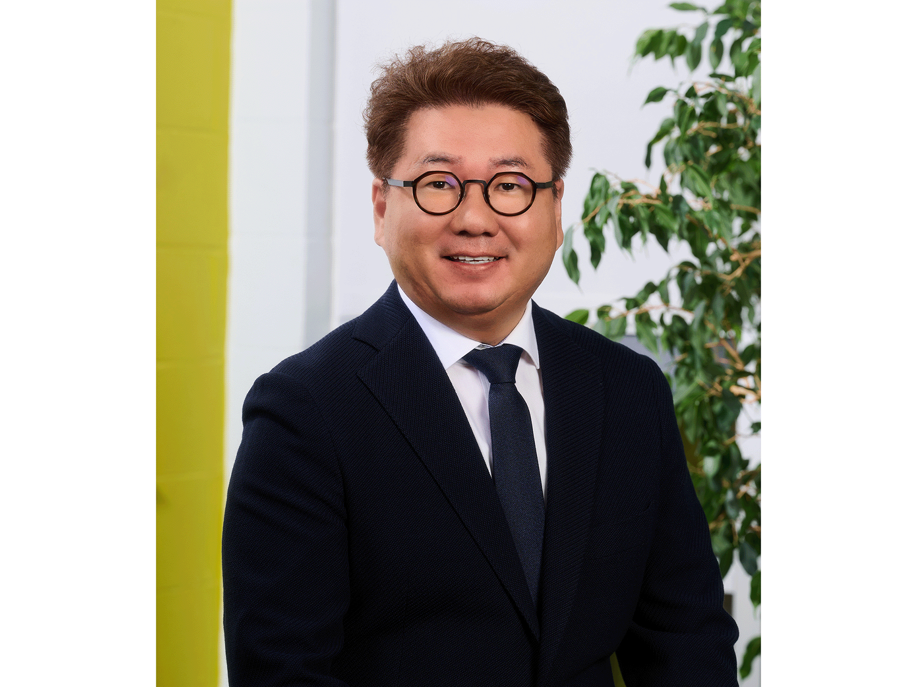 Portrait of Soo-yong Byun wearing black suit and tie and black, round-rimmed glasses.