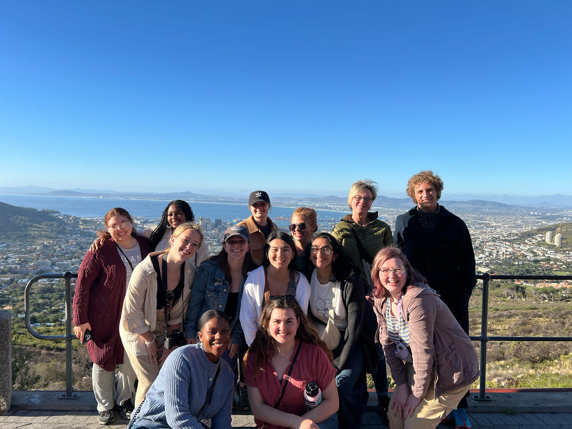 Students stand on top of a mountain with Cape Town, South Africa, in the distance behind them. From left to right, back row: Monica Rodriguez, Safian Lewis, Corinne Chase, Jill Wood, Susanne M. Klausen, Ethan Capitano. Middle row: Payton Smith, Cara Arnoldi, Samantha Powell, Keya Ahrestani, Hannah Yurack. Front row: Phathutshedzo Bale (local staff member), Maggie Day