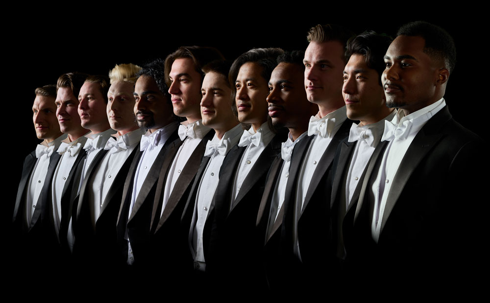 A group twelve diverse me wearing tuxedos stand and look in the same direction.