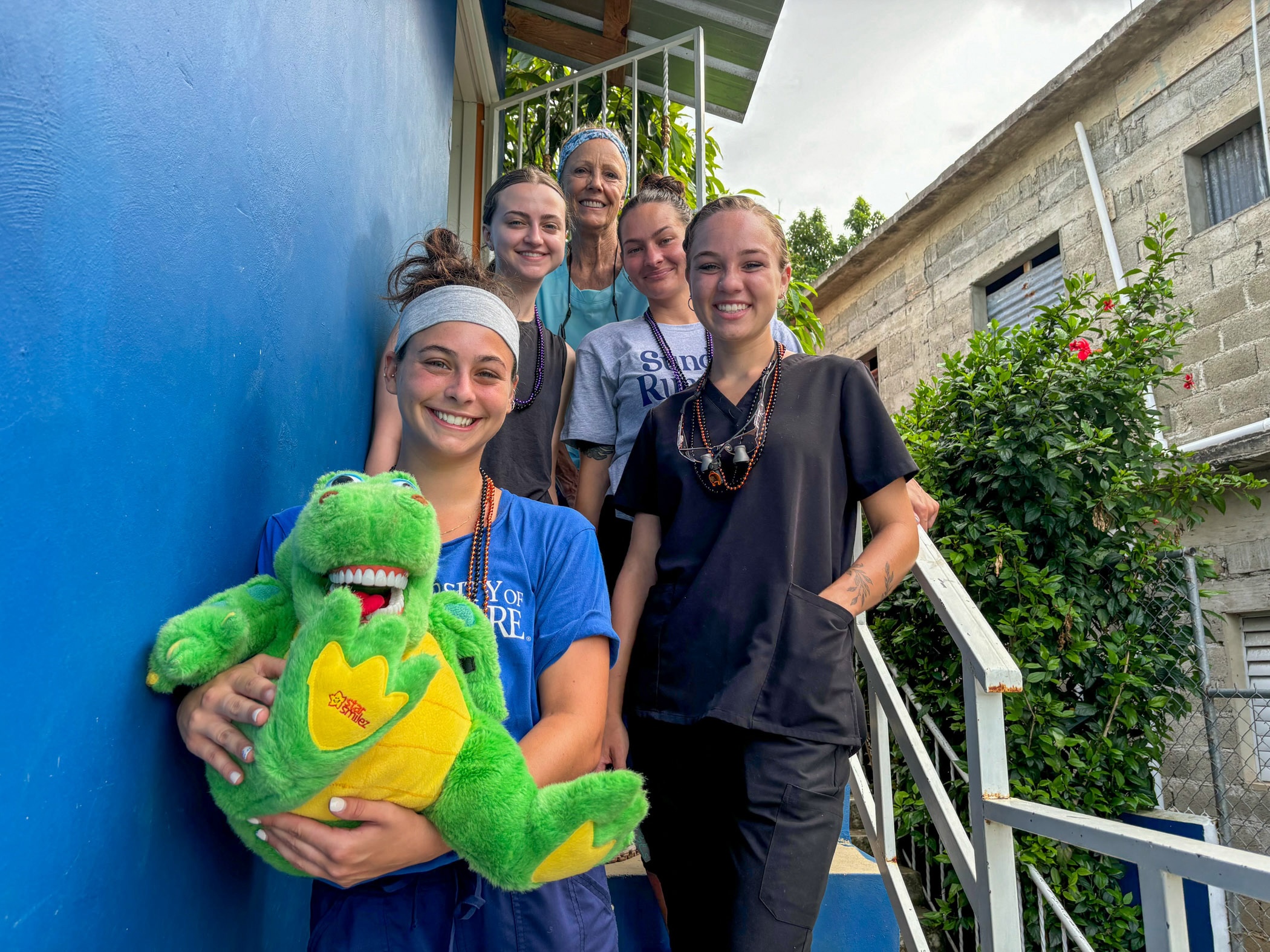 Dental hygiene students together on steps with a blue wall beside them. One of them is holding a green and yellow stuffed animal.