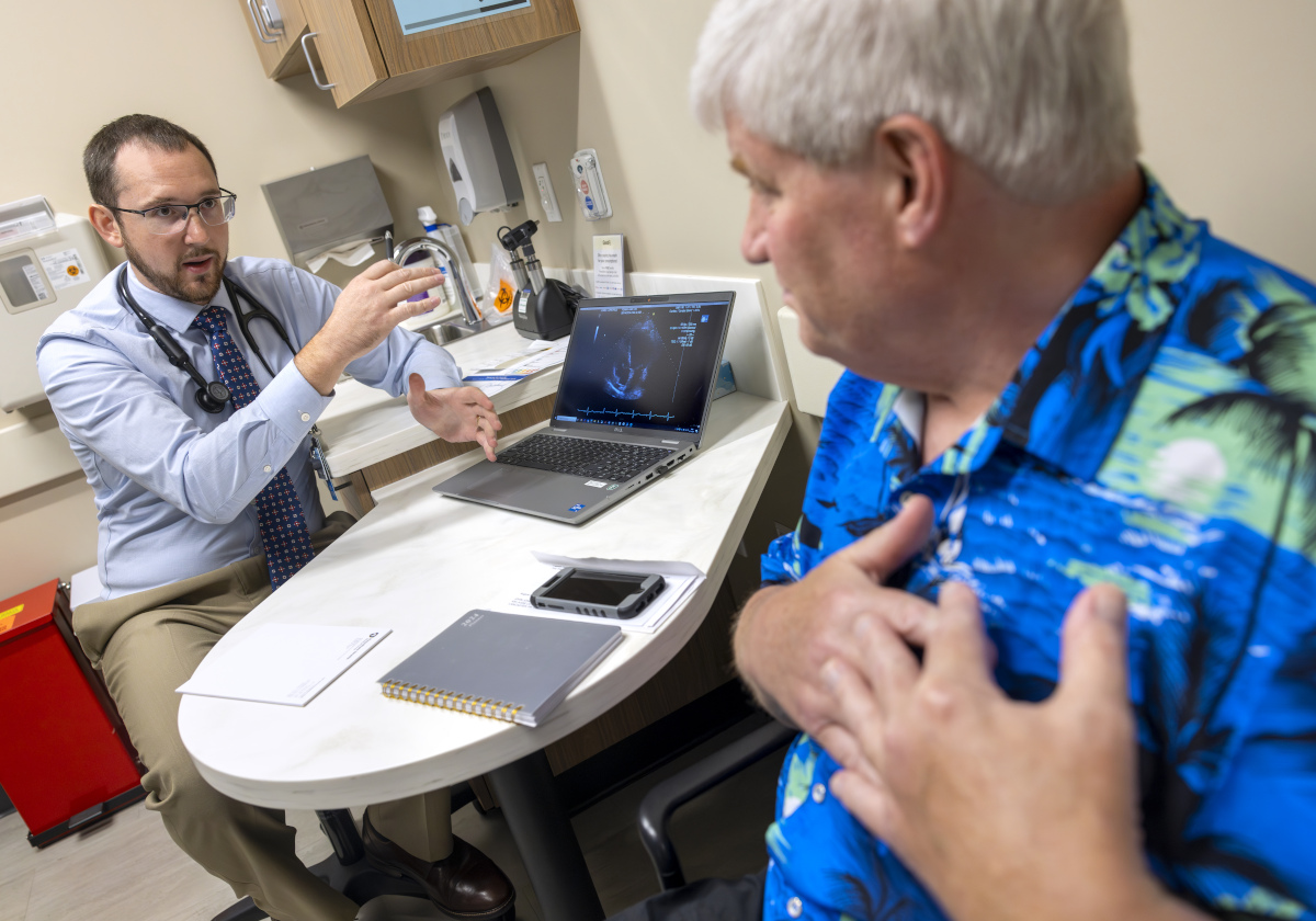 Tyler Thomas, left, gestures as he talks to patient John Jones, right. Thomas is wearing a button-down shirt, tie, glasses and has a stethoscope around his neck. Jones is wearing a Hawaiian shirt and has hands over his heart.