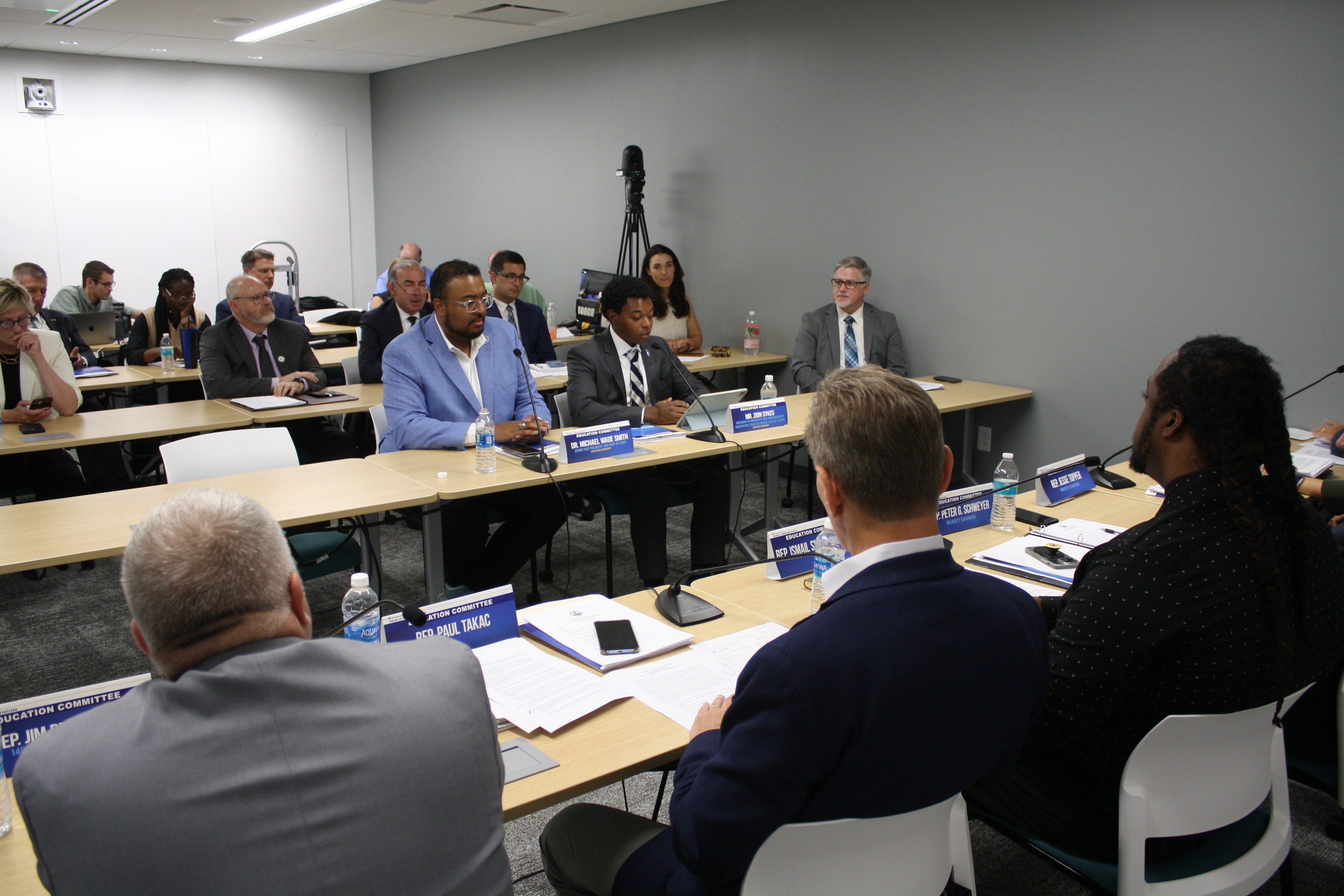 two men sitting behind a table infront of microphones infront of them speaking to a room full of people