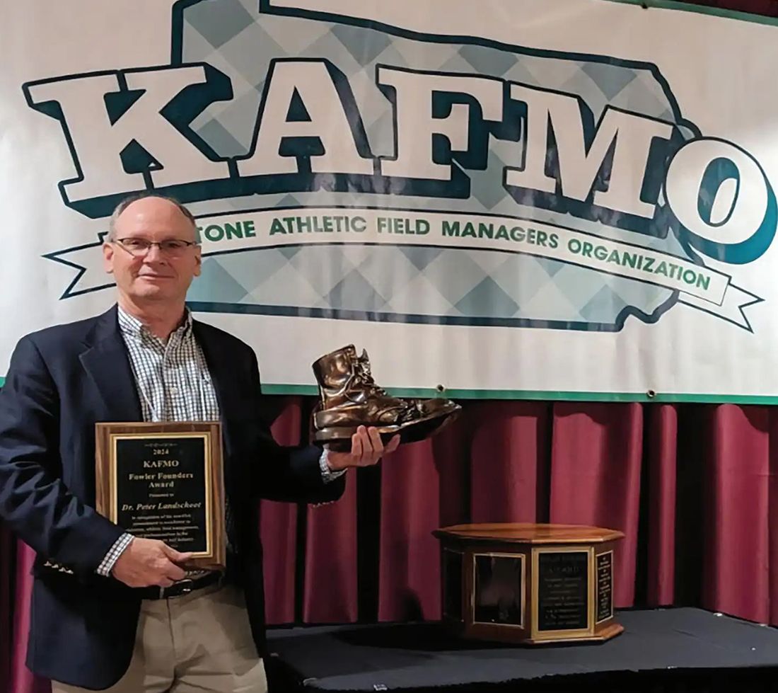 Person standing with a plaque and a bronze shoe in front of a sign reading "KAFMO"