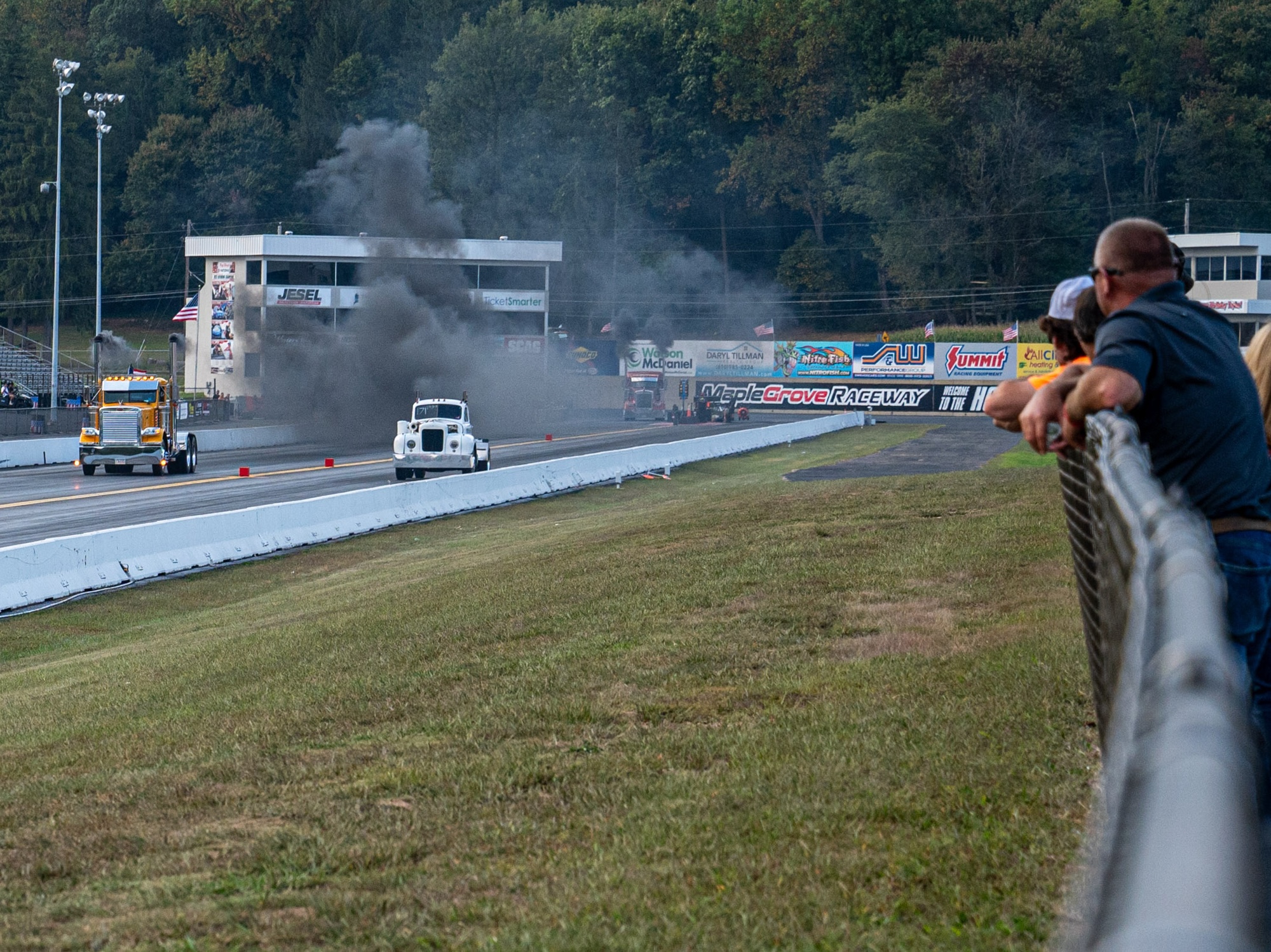 Two trucks speed down a drag strip as members of the crowd watch along the fence.