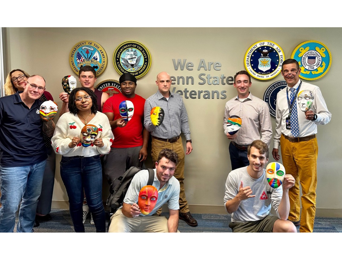 Penn State student veterans hold up masks they created during their first-year seminar. Behind them there is a sign on the wall that says "We Are Penn State Veterans."