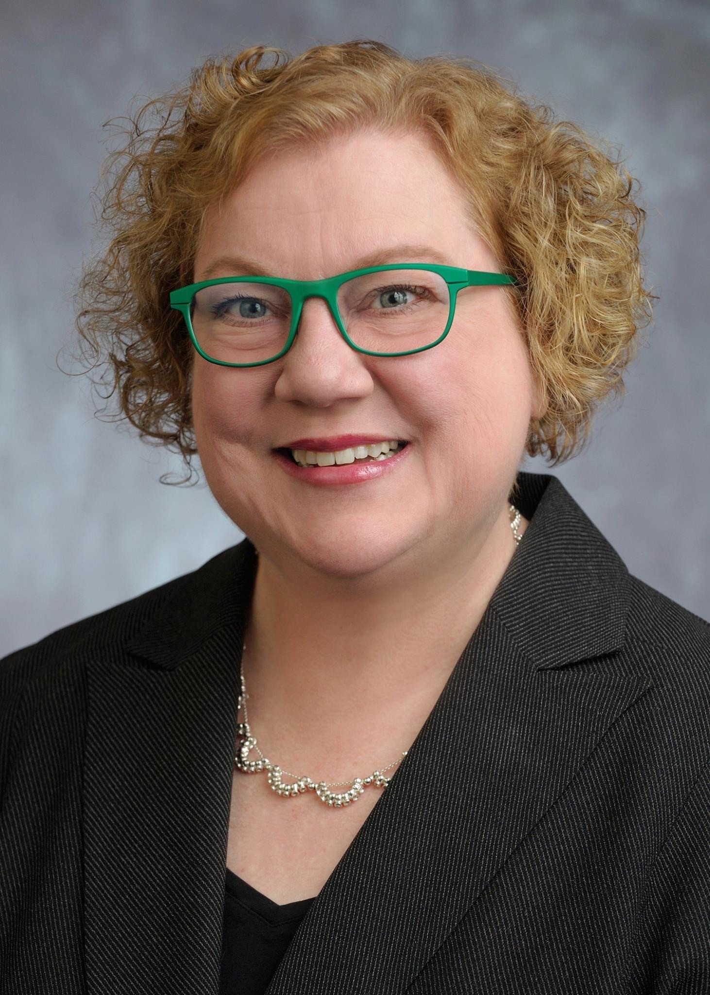 Head shot of a woman with blonde curly hair and green-rimmed glasses smilng