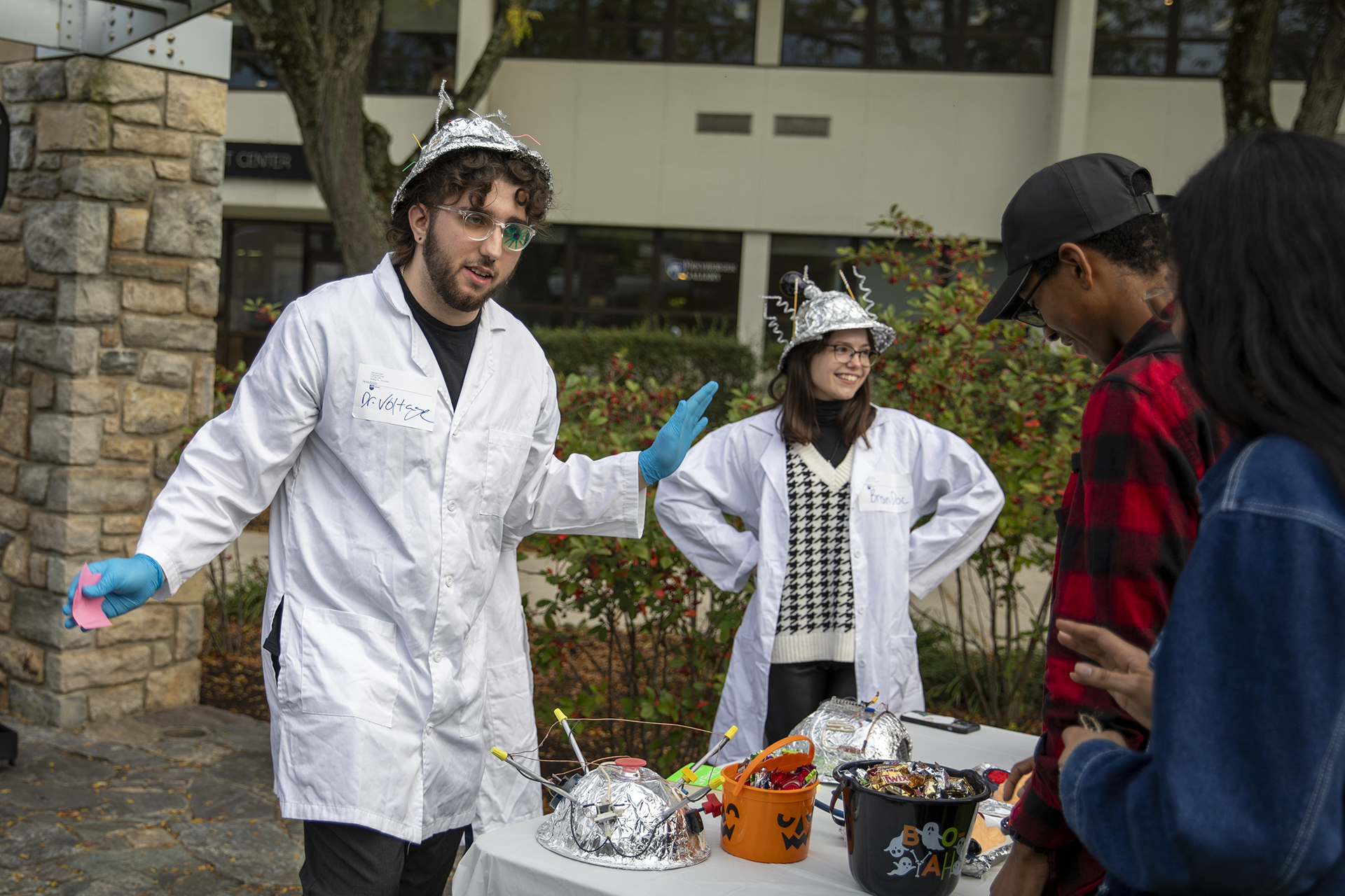 Students wearing lab coats and tinfoil hats conduct experiments with onlookers