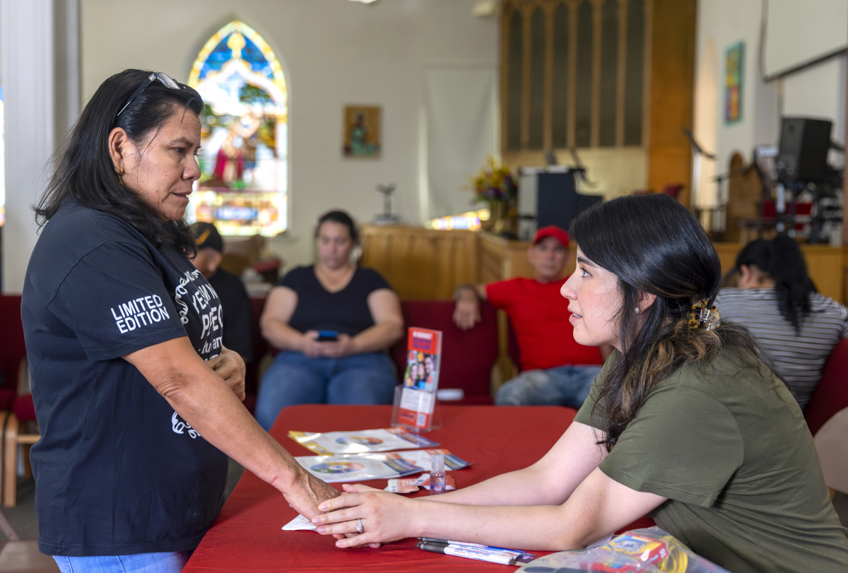 Two women hold hands as they speak to one another. One woman sits at a table and the other stands. They’re in a church, a stained glass window is behind them and people in a pew look on.